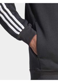 Μαύρη φόρμα με λευκές ρίγες στα μανίκια, που διαθέτει τσέπη στη μέση, φτιαγμένη από μαλακό, υφασματώδη υλικό.