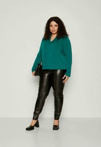 Türkiser, langärmeliger V-Ausschnitt-Pullover kombiniert mit schwarzen, fauxledernen Hosen. Das Model hält eine schwarze Handtasche und trägt schlichte schwarze Slippers.