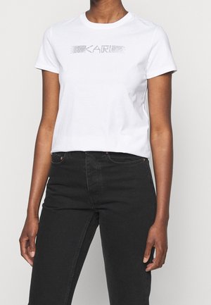 Osoba w białej koszulce z krótkim rękawem z napisem „KARL” utworzonym z kropek, w połączeniu z czarnymi, wysokim stanem jeansami.