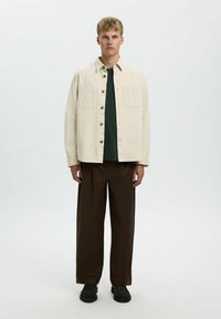 Chaqueta de camisa beige claro con dos bolsillos en el pecho, superpuesta a una camisa verde oscuro, combinada con pantalones sueltos marrón oscuro y zapatos negros.