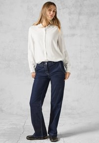 Chemise blanche à manches longues avec des boutons, associée à un jean large bleu foncé avec des surpiqûres contrastantes et des chaussures noires plates ornées de clous.
