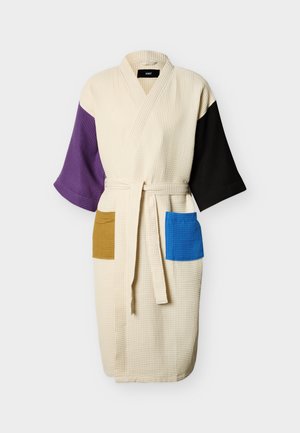 Albornoz color crema con textura de waffle, manga izquierda morada, manga derecha negra, bolsillo izquierdo mostaza y bolsillo derecho azul, atado con un cinturón.