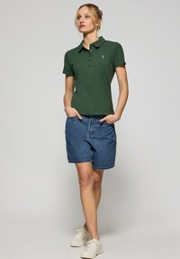 Zielona koszulka polo z kołnierzykiem i trzema guzikami, zestawiona z niebieskimi szortami dżinsowymi. Model nosi białe trampki, prezentując swobodny strój.