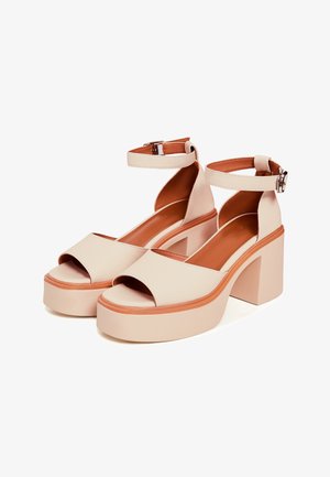 Mujer con sandalias beige de plataforma y tacón bloque con tiras en el tobillo, sentada en una silla de madera, ajustando una de las tiras de la sandalia.
