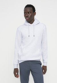 Νεαρός άνδρας που φοράει ανοιχτό γκρι φουτεράκι με κουκούλα και σκουρόχρωμο γκρι παντελόνι, στέκεται με τα χέρια χαλαρά, κοιτάζοντας ελαφρώς προς τα πλάγια με φόντο έναν απλό τοίχο.