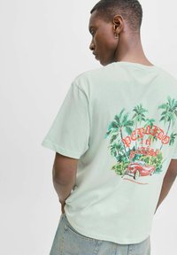 Miesta zaļas kokvilnas T-krekls, kas aizmugurē rotāts ar krāsainu grafiku ar palmu kokiem un vintage automašīnu, un tekstu "Bienvenidos a Puerto Rico".