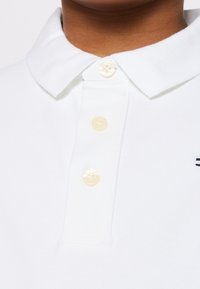 Gros plan d'une personne portant un polo blanc à col avec trois boutons crème et une broderie sombre partielle sur le côté droit.