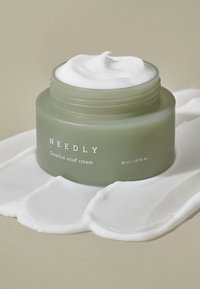 Tarro verde esmerilado de crema para alivio de cicatrices con una textura blanca y cremosa como nata montada. Etiqueta visible con la palabra "NEEDLY", reposando sobre un charco de crema blanca.