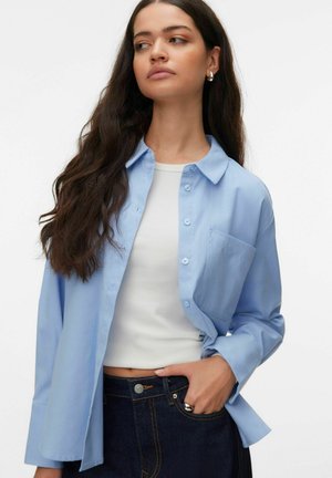 Gaiši zils pogās aiztaisāms krekls ar apkakli, garām piedurknēm un vienu krūšu kabatu, valkāts pār baltu topu un kombinēts ar tumši zilām džinsām.