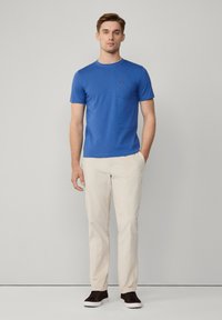 Niebieska bawełniana koszulka z okrągłym dekoltem i kieszonką na piersi, zestawiona z jasnobeżowymi spodniami i brązowymi trampkami. Prosty i casualowy styl.