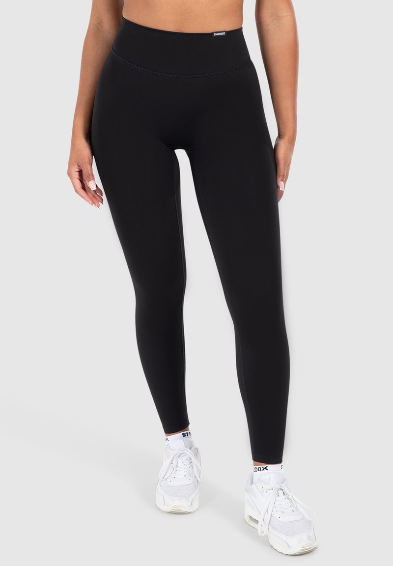 Leggings neri a vita alta realizzati in materiale elasticizzato, con una superficie liscia e un design elegante, abbinati a scarpe sportive bianche.