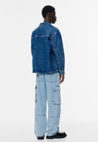 Denim jakna v temno modri barvi, ki ima klasičen ovratnik in šivane detajle, v kombinaciji s svetlo modrimi cargo hlačami z bočnimi žepi.