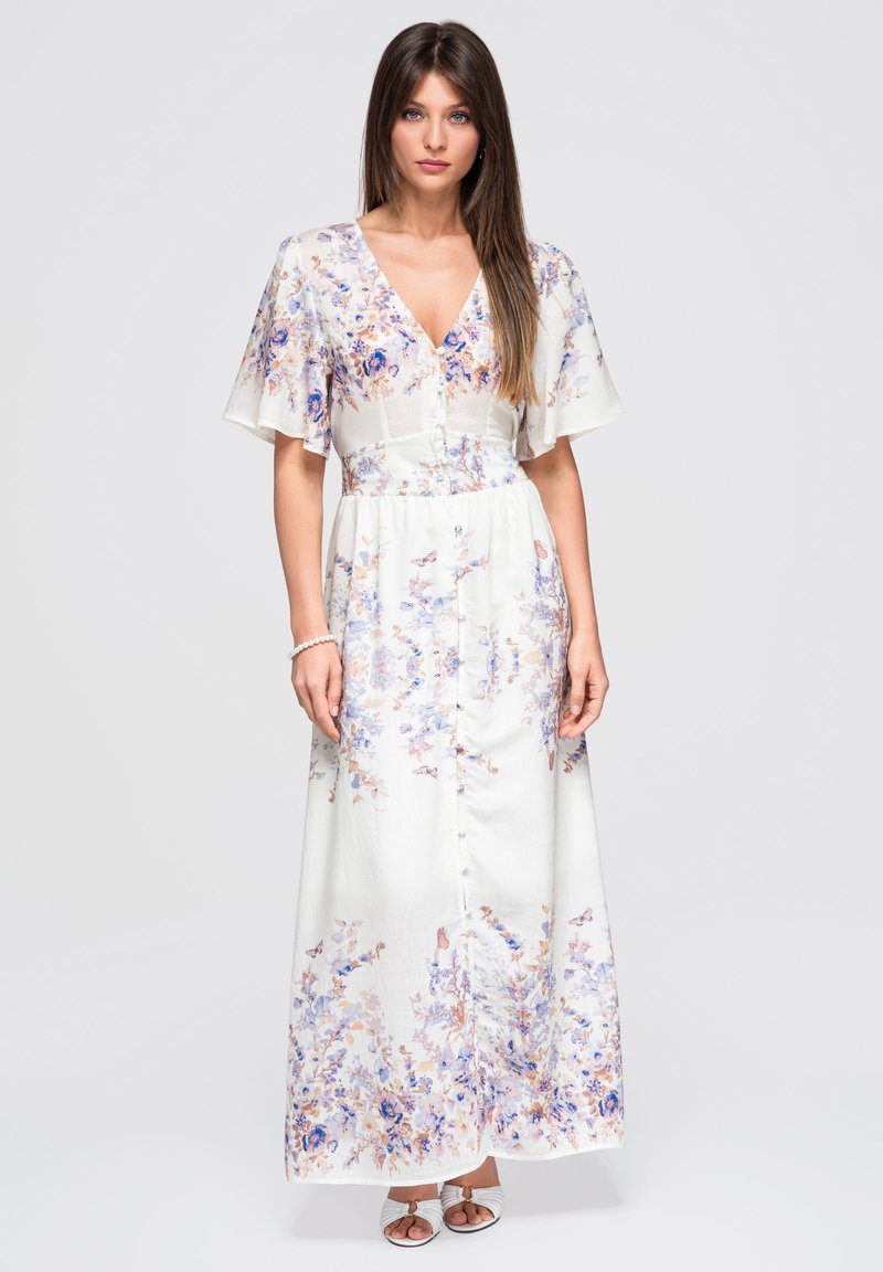 Jeune femme portant une robe longue blanche à fleurs, avec de courtes manches volantées, un décolleté en V, des boutons sur le devant et des sandales blanches à bouts ouverts.