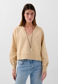 Beige V-Ausschnitt Sweatshirt mit leicht taillierter Silhouette, gerippten Bündchen und strukturiertem Stoff; kombiniert mit hellblauen Jeans und einer runden Halskette.