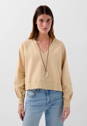 Beige V-hals sweatshirt met een licht getailleerde silhouet, geribbelde manchetten en een gestructureerde stof; gecombineerd met lichtblauwe jeans en een ronde ketting.