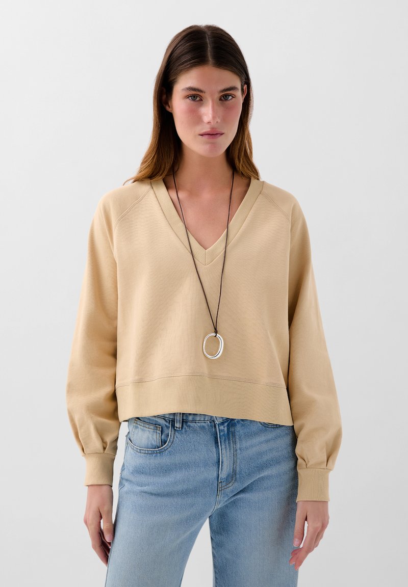 Beige V-Ausschnitt Sweatshirt mit leicht taillierter Silhouette, gerippten Bündchen und strukturiertem Stoff; kombiniert mit hellblauen Jeans und einer runden Halskette.