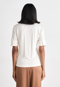 Lysbeige ribbet topp med korte ermer, med rullet mansjett og en tettsittende silhuett, laget av et teksturert stoff.