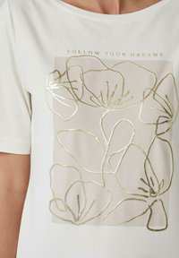 T-shirt blanc avec un décolleté plongeant, présentant un design floral en feuille d'or et le texte "SUIVEZ VOS RÊVES" au-dessus du motif.