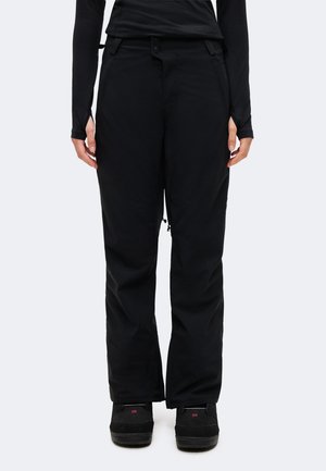 Une personne présente un pantalon noir ample à taille boutonnée et des chaussures noires sur un fond clair et uni.