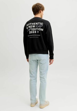 Man draagt zwarte trui met witte tekst "Authentic & New Edition 2023" op de rug, lichtblauwe spijkerbroek en beige sneakers, staand met de rug naar ons toe.