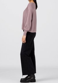Pull oversized lilas avec des manches bouffantes, associé à un jean noir taille haute à jambes larges et des bottes noires à lacets. Fond uni.
