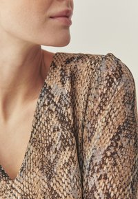 Braun-beiges Schlangenprint-Stoff auf einem V-Ausschnitt-Top, mit einem leichten, strukturierten Gefühl und sanftem Fall um die Schultern.