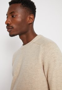 Beige genser med teksturert overflaten, rund hals, og raglanermer, vist mot en ensfarget bakgrunn.