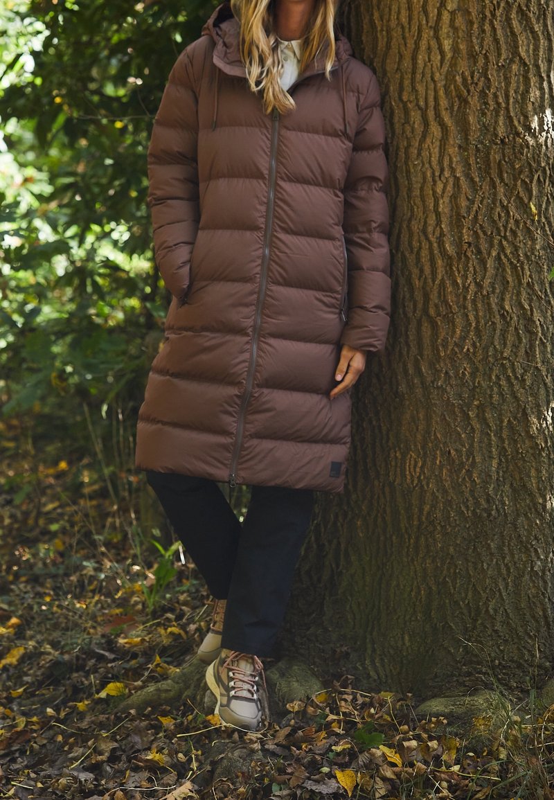 Brun puffjacka med en strukturerad yta och huva, med en framdragkedja och sidofickor, i kombination med mörka byxor och neutrala skor.