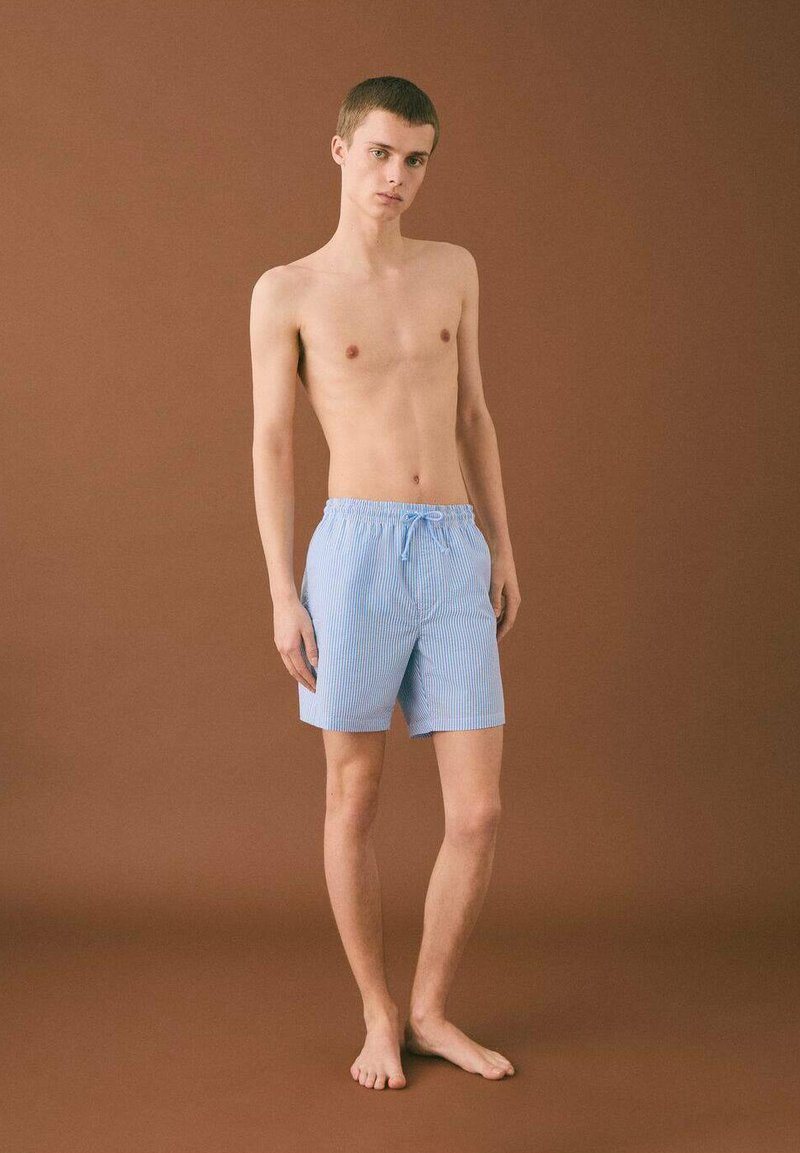 Modra in bela prugasta kopalna kratka hlača z elastičnim pasom in vrvico. Nosijo jih model na nevtralni rjavem ozadju.
