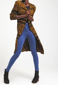 Lång stickad cardigan i svart och guld mönster, med öppen front och trekvartslånga ärmar; kombinerad med blå skinny jeans och svarta ankelboots.