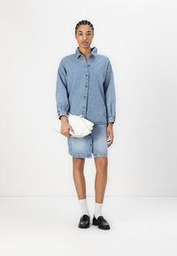 Denim džinsinė striukė su mygtukais, mėlynos spalvos, derinama su atitinkamu džinsiniu sijonu. Balta rankinė ir juodi mokasīnai papildo aprangą.