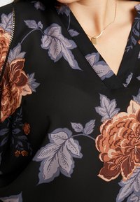 Blusa nera con grandi motivi floreali in arancione, bordeaux e blu chiaro. Design a V con tessuto morbido e leggero.