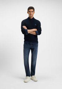 Suéter azul marino con cuello alto, tejido texturizado, combinado con jeans azul oscuro y zapatillas blancas. El modelo está de pie con los brazos cruzados.