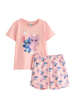 Ung pige i pink T-shirt og shorts, begge med farverige tegneseriefigurer, stående barfodet mod en ensfarvet baggrund.