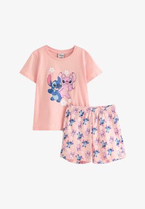 Ung pige i pink T-shirt og shorts, begge med farverige tegneseriefigurer, stående barfodet mod en ensfarvet baggrund.