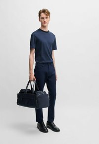 Maglietta blu navy con colletto tondo, pantaloni scuri e scarpe nere. Porta una borsa a tracolla blu navy con texture, due manici e cerniere.