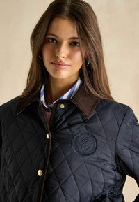 Pikowana ciemnoniebieska kurtka z brązowym kołnierzem z korduroju, złotymi akcentami i wytłoczonym logo. Noszona na paskowanej koszuli o teksturowanym wykończeniu.