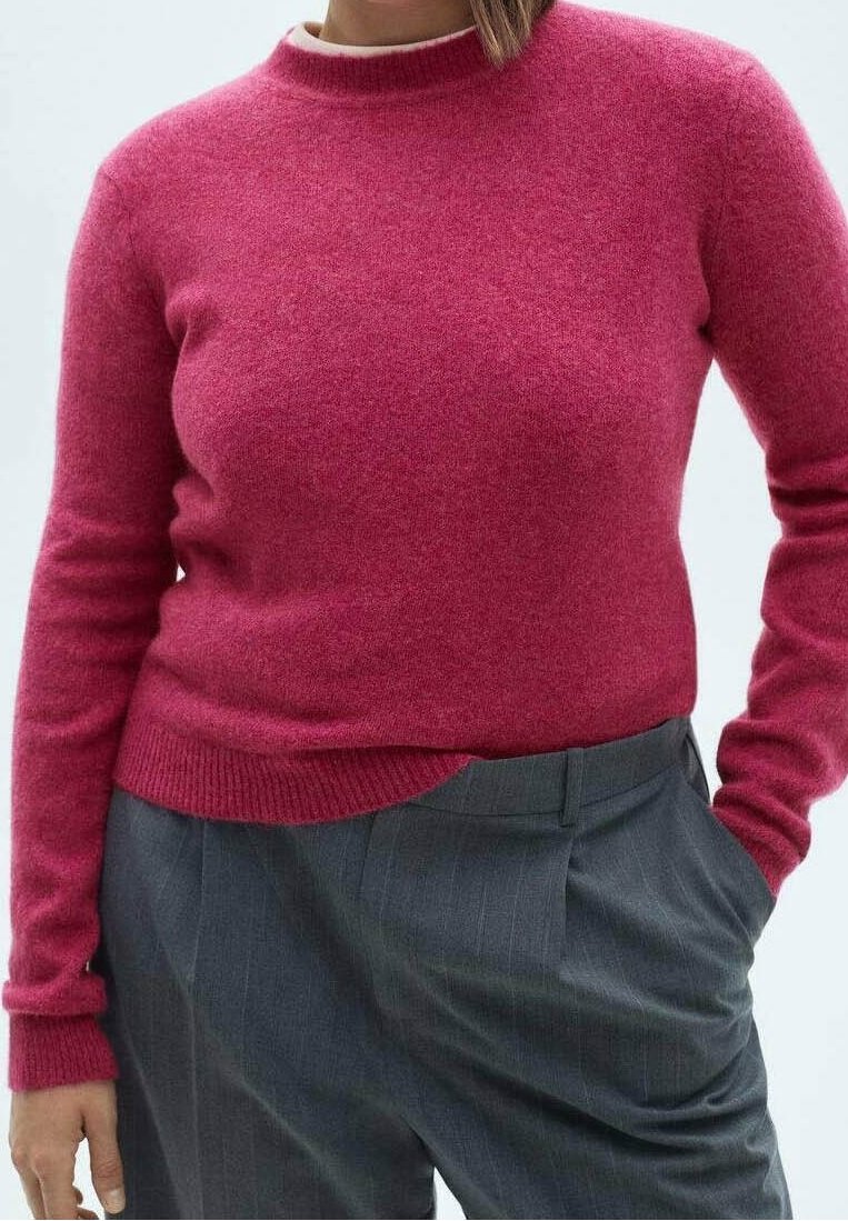 Personne portant un pull à manches longues rose vif rentré dans un pantalon taille haute gris à fines rayures, une main dans la poche.