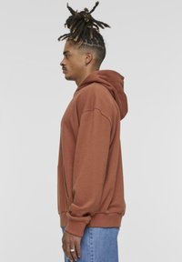 Sudadera con capucha marrón de corte holgado, con bolsillo tipo canguro y puños acanalados, combinada con jeans de color azul claro. Silueta casual y relajada.
