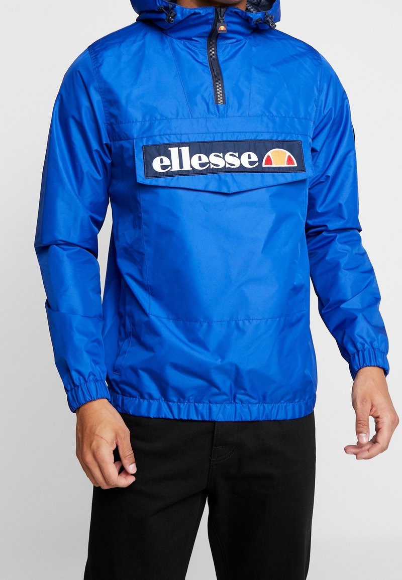 Man som bär en ljusblå tröja med ficka fram och "ellesse"-logotyp, tillsammans med svarta byxor.