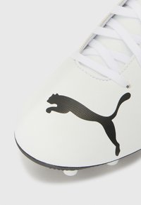 Crampons de football blancs avec une tige synthétique lisse, présentant un logo Puma noir bien visible, des lacets blancs et une semelle extérieure noire avec des crampons.