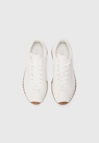 White sneakers, smooth synthetic upper, round toe, flat laces, and brown rubber outsole with textured pattern. Minimalist design.