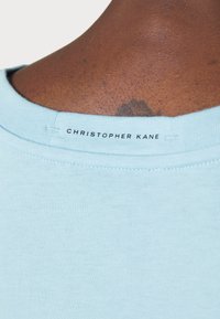 Hellblaue Baumwoll-T-Shirt, Rundhalsausschnitt, mit schwarzem "CHRISTOPHER KANE"-Label an der hinteren Ausschnittkante gestickt; glatte, weiche Textur.
