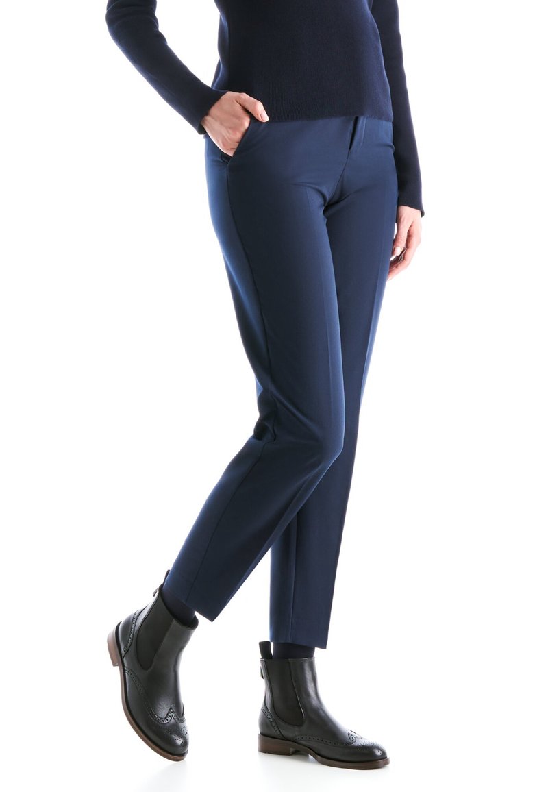 Femme portant un pantalon slim bleu marine, des bottines noires et un haut bleu marine à manches longues, debout avec une main dans la poche.