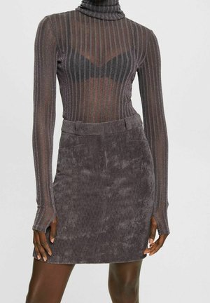 Vrouw draagt een donkergrijs geribbeld coltrui gaas topje over een zwarte beha en een hoog taille grijze corduroy minirok.