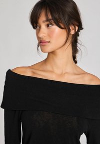 Juodas off-the-shoulder top'as, pagamintas iš minkštos megztos medžiagos, su plačiu apykaklės iškirpimu ir ilgomis rankovėmis, šiek tiek skaidraus tekstūros.
