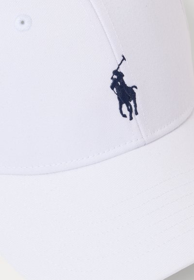 Balta beisbolo kepurė, pagaminta iš medvilnės, su tamsiai mėlyna siuvinėta polo žaidėjo logo kairėje pusėje, struktūrizuota kraštelio forma ir siūlėmis detalėse.