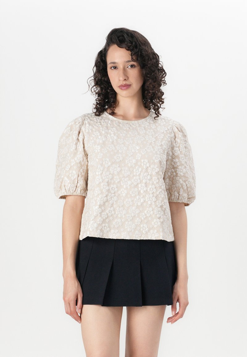 Lys beige bluse med teksturert blomstermønster, puffede kort ermer og rund halslinning, kombinert med et svart plisserte skjørt.