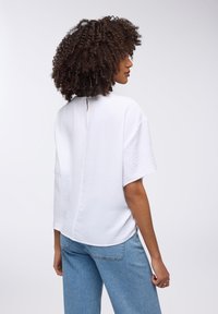 Top bianco a maniche corte con superficie texture, caratterizzato da un'apertura a chiave sul retro e una leggera caduta. Abbinato a jeans azzurri chiari.