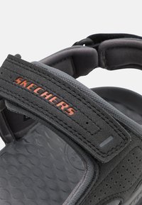 Svarta Skechers-sandaler med justerbar rem, gummisula, texturerad fotbädd och kontrasterande orange logotyp på remmen, med vadderat stöd.